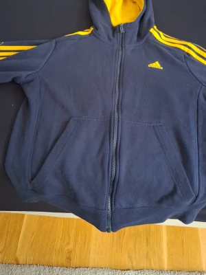 Mörkblå Adidas hoodie med gula detaljer - Snygg mörkblå hoodie från Adidas med dragkedja, gula ränder på ärmarna och gul insida i huvan. Klassisk Adidas-logga på bröstet och två fickor framtill. Perfekt för en sportig och avslappnad stil.