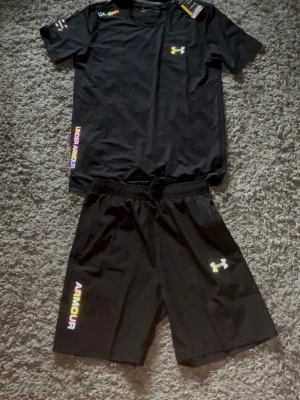 Svart träningsset från Under Armour - Snyggt svart träningsset från Under Armour med t-shirt och shorts. Setet har färgglada logotyper och textdetaljer på både tröja och byxa. Tillverkat i ett lätt och snabbtorkande material som passar perfekt till träning eller sport.