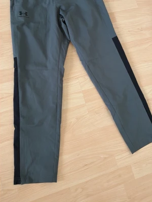 Under Armour byxor  - Snygga grå träningsbyxor från Under Armour med svarta detaljer längs sidorna. Byxorna har en rak passform och är tillverkade i ett lätt och funktionellt material som passar perfekt för träning eller sportaktiviteter.