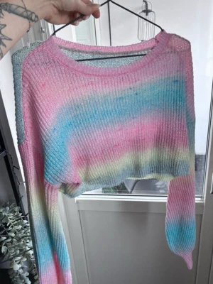 Pastellfärgad cropped stickad tröja - Säljer en stickad cropped tröja i pastellfärger med inslag av rosa, blått, gult och grönt. Tröjan har rund halsringning, långa ärmar och ett ribbstickat mönster. Perfekt för dig som gillar färgglada och unika plagg.
