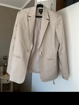 Beige kavaj från Boohoo - Stilren beige kavaj från Boohoo med klassisk passform och en knapp framtill. Kavajen har två fickor utan lock och är helfodrad. Perfekt för att lyfta vilken outfit som helst med en clean och modern look.