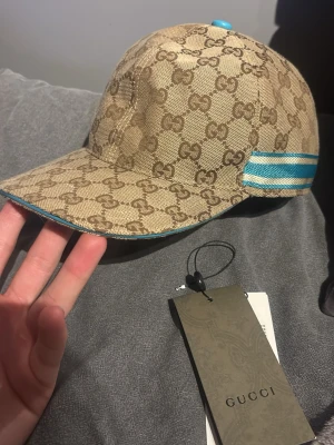 GG keps beige - Snygg beige keps från GG med klassiskt GG-mönster över hela kepsen. Kepsen har blå och vita ränder på sidan samt blå kant runt skärmen. Tillverkad i canvasmaterial och har en böjd skärm för en sportig look.