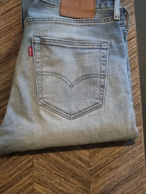 Ljusblå slitna Levi's jeans 34 34  - Säljer ett par ljusblå Levi's jeans med klassisk femficksdesign och en snyggt sliten look. Jeansen har en trasig detalj vid knät och är tillverkade i bomull. Perfekta för dig som gillar en avslappnad och trendig stil.