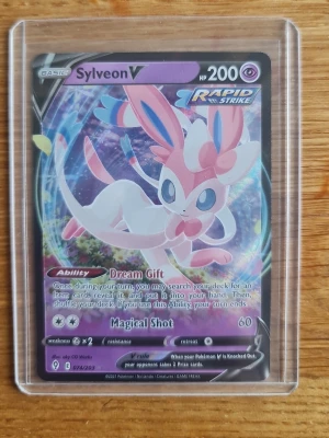 Sylveon pokemon card  - Unikt Pokémonkort med Sylveon V Rapid Strike, HP 200. Kortet har färger i vitt, rosa och blått, och är tillverkat i laminerad papp. Det är förvarat i en transparent plastficka och har holografiska detaljer. Perfekt för samlare eller Pokémon-fans.