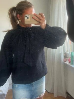 Svart blus med knytband i halsen - Trendig svart blus med voluminösa långärmar och snyggt knytband i halsen. Blusen har en rak passform och är tillverkad i ett lätt, vävt tyg som ger en elegant look. Perfekt att styla med jeans eller kjol för en cool outfit.