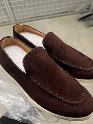 Bruna mocka loafers med vit sula - Snygga loafers i mörkbrun mocka med kontrasterande vit sula. Skorna har en klassisk slip-on design och diskreta sömmar längs ovandelen. Insidan är fodrad i beige och de har en stilren, minimalistisk look som passar till många outfits.