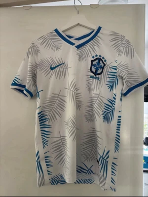 Vit Brasilien fotbollströja Nike - Snygg vit fotbollströja från Nike med Brasilien CBF-logga på bröstet. Tröjan har blå och grå palmbladsmönster, blå detaljer vid krage och ärmslut samt rund halsringning. Perfekt för dig som älskar fotboll och vill sticka ut på planen.