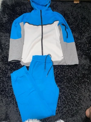 Blå och grå loungewear set med huva - Snyggt loungewear set med zip-hoodie och matchande byxor. Tröjan har blå huva och axlar, vita partier framtill och grå ärmar. Byxorna är helblå med dragsko i midjan. Perfekt för chill dagar hemma eller när du vill vara bekväm.