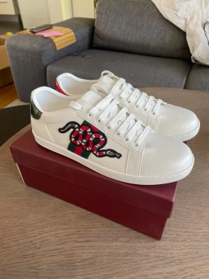 Gucci vita sneakers med orm - Snygga vita sneakers från Gucci med broderad röd och svart orm på sidan och gröna/röda detaljer. Skorna har klassisk låg profil, vita snören och är tillverkade i läder. Perfekta för dig som vill sticka ut med en lyxig och trendig look.