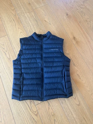 Marinblå dunväst från J.Lindeberg - Snygg marinblå dunväst från J.Lindeberg med quiltad design och diskret logga på bröstet. Västen har dragkedja framtill och två sidofickor. Perfekt för lager-på-lager och ger en sportig look. Tillverkad i lätt och värmande material. Den är liten i storlek så den sitter lite mellan en S och M