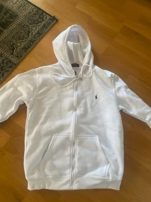 Vit hoodie från Polo Ralph Lauren - Säljer en vit hoodie från Polo Ralph Lauren med dragkedja framtill och klassisk blå logga broderad på bröstet. Hoodien har huva med dragsko och två stora fickor framtill. Tillverkad i mjuk bomullsblandning för skön känsla.