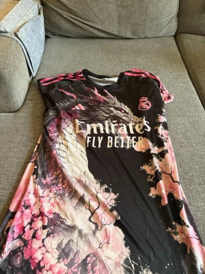 Real Madrid x Adidas draktröja L - Cool Real Madrid-tröja från Adidas i storlek L med unikt drakmotiv och körsbärsblommor i rosa och vitt på svart bakgrund. Tröjan har rosa detaljer på axlarna och Aeroready-material som andas. Perfekt för dig som vill sticka ut på planen.