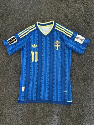 Blå Sverige fotbollströja  - Säljer en blå fotbollströja från Adidas med Sveriges landslagsemblem på bröstet och nummer 11. Tröjan har gula detaljer, mönstrad design och klassiska Adidas-ränder på axlarna. Perfekt för dig som älskar fotboll och vill visa ditt stöd för Sverige. En replika