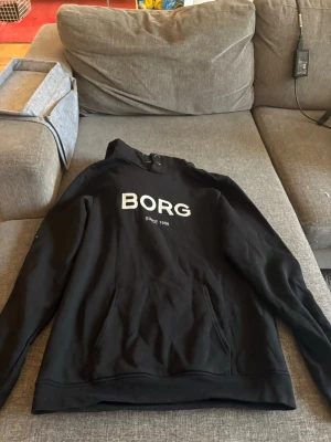 Svart hoodie från Björn Borg - Snygg svart hoodie från Björn Borg med stor vit BORG-logga på bröstet och texten 'Since 1984' under. Klassisk känguruficka framtill och huva utan dragsko. Perfekt för en chill och sportig look.