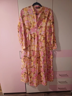 Klänning - Långklänning från LCW Modest med färgglatt blommigt mönster i rosa, gul, beige och brunt. Klänningen har knappar framtill, lång ärm och en avslappnad passform. Perfekt för dig som gillar färg och mönster!