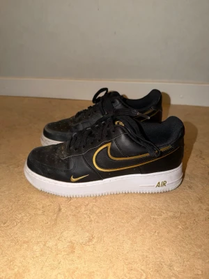 Nike Air Force 1 svart/guld sneakers - Snygga Nike Air Force 1 sneakers i svart läder med guldfärgade detaljer och vit sula. Klassisk låg modell med svart snörning och ikonisk Swoosh-logga i guld. Perfekta för dig som vill ha en stilren och sportig look.