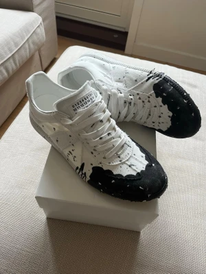 Maison Margiela svartvita sneakers - Storlek 46/ passar 45. Unika sneakers från Maison Margiela i vitt med svart färgstänk och snörning. Skorna har en cool, konstnärlig look med svart detalj på tå och sidor. Märkets ikoniska etikett syns på plösen och insidan. Perfekta för dig som gillar statement-plagg.