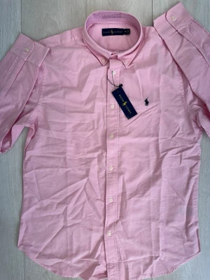Rosa skjorta från Ralph Lauren - Snygg rosa skjorta från Ralph Lauren med klassisk button-down krage och broderad logga på bröstet. Skjortan har korta ärmar och knäppning framtill. Perfekt för en stilren look och tillverkad i mjuk bomull.