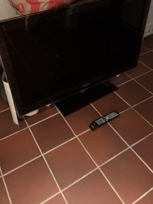Svart platt-TV med fjärrkontroll - Säljer en svart platt-TV med tillhörande fjärrkontroll. TV:n har en stilren, rektangulär design och passar perfekt för filmkvällar eller gaming. Fjärrkontrollen har många knappar för olika funktioner. Märke och exakt storlek är okänd.