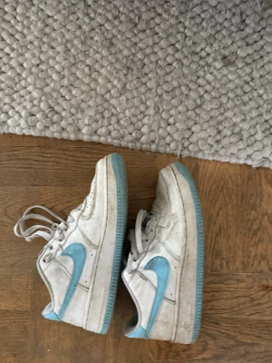 Nike vita sneakers med blå swoosh - Säljer ett par klassiska vita Nike sneakers med ljusblå swoosh och matchande blå yttersula. Skorna har lågt skaft, vita skosnören och är tillverkade i läder. Perfekta för dig som gillar retrostil och streetwear.