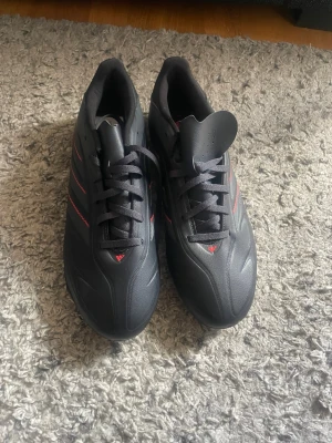 Svarta Adidas fotbollsskor med dobbar - Säljer ett par svarta Adidas fotbollsskor med röda detaljer och klassiska tre ränder på sidan. Skorna har snörning och är tillverkade i syntetmaterial med dobbar för spel på gräs. Perfekta för dig som vill ha stilrena och funktionella skor på planen.