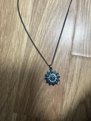 Halsband silver/blå - Säljer ett halsband med en blomformad berlock dekorerad med blå pärlor och små stenar. Kedjan är silverfärgad och tunn, vilket ger ett stilrent intryck. Perfekt för dig som gillar unika accessoarer med färg och detaljer.