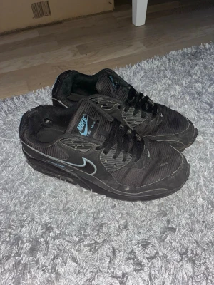 Nike Air Max 90 svarta sneakers - Svarta Nike Air Max 90 sneakers med mesh och syntetmaterial. Skorna har klassisk Nike-logga i ljusblått på tungan och sidan, samt svarta skosnören och sula. Perfekt för dig som gillar sportig och stilren look.
