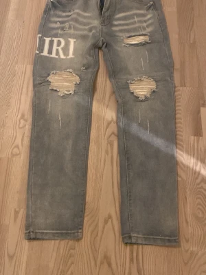 Grå slitna jeans med tryck - Säljer ett par grå jeans med stora slitningar på båda benen och vita bokstavstryck på framsidan och baksidan. Jeansen har rak passform och coolt distressed utseende som sticker ut. De är XL