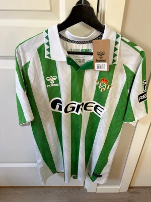 Grönvit fotbollströja Real Betis Anthony  - Säljer en grön och vit randig fotbollströja från Real Betis, tillverkad av Hummel. Tröjan har klassisk krage, klubbens emblem på bröstet och sponsorloggor. På ryggen står det 'ANTONY' och nummer 7. Perfekt för dig som älskar fotboll och vill sticka ut på planen.