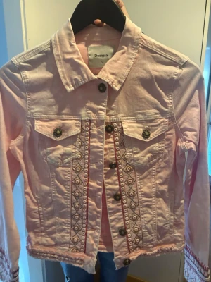 Ljusrosa jeansjacka från Desigual - Ljusrosa jeansjacka från Desigual med snygga broderade detaljer längs knappraden och på ärmarna. Jackan har klassisk krage, två bröstfickor med knappar och dekorativa sömmar. Perfekt för dig som gillar unika plagg med coola mönster.