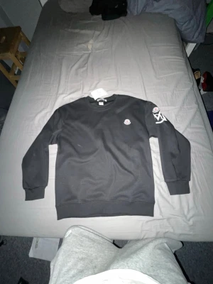 Svart Moncler sweatshirt med logga - Svart sweatshirt från Moncler med rund halsringning och ribbade muddar. Liten Moncler-logga på bröstet och en större vit detalj på ena ärmen. Perfekt för en stilren och avslappnad look.