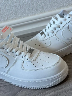 Nike Air Force 1 vita sneakers - Klassiska Nike Air Force 1 sneakers i helvitt läder med perforerad tå och ikonisk Swoosh på sidan. Skorna har vita skosnören, robust yttersula och låg profil som ger en clean och tidlös look. Perfekta för dig som vill ha en stilren sneaker till din outfit.