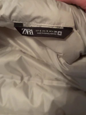 Beige/vit dunväst från Zara - Snygg beige dunväst från Zara i storlek S. Västen har quiltad design och är tillverkad i ett glansigt material som ger en modern look. Perfekt att slänga över en hoodie eller tröja för extra värme. Dragkedja framtill och diskret logotyp på insidan.