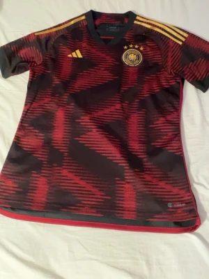 Fotbollströja - Säljer en officiell Tyskland matchtröja från Adidas i rött och svart med gula detaljer. Tröjan har korta ärmar, V-ringning och tre stjärnor ovanför landslagets emblem. Tillverkad i lätt och ventilerande material, perfekt för fotboll eller träning.
