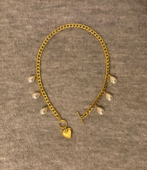 Guldhalsband med pärldroppar och hjärta - Snyggt halsband i guldfärgad metall med fem vita pärldroppar och ett litet hjärthänge. Kedjan har en chunky look och stängs med en toggle-lås. Perfekt för dig som vill ha något som sticker ut lite extra.