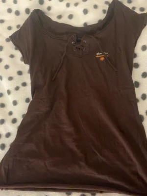 Brun topp med snörning från Atom Girl - Cool brun topp från Atom Girl med snörning framtill och broderad logga på bröstet. T-shirten har korta ärmar och en avslappnad passform. Perfekt för dig som gillar en enkel men unik stil.