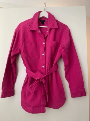 Denim skjorta från Lindex - Stilren ceriserosa overshirt från Lindex med knytskärp i midjan, tryckknappar framtill och två bröstfickor. Tillverkad i ett mjukt bomullsliknande material och har klassisk krage samt långa ärmar. Perfekt för lager på lager och ger en färgklick till din outfit.