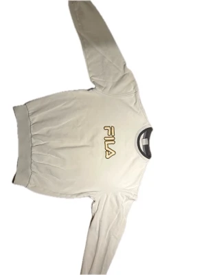Vit sweatshirt från FILA - Säljer en vit sweatshirt från FILA med broderad logga i gult och svart på bröstet. Tröjan har rund halsringning med kontrastfärg, ribbade muddar vid ärmslut och nederkant. Perfekt för en sportig och avslappnad stil.