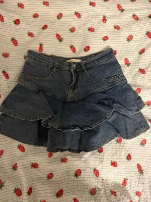 Blå volangkjol i denim - Säljer en blå jeanskjol med volangdetaljer och klassiska fickor fram och bak. Kjolen har en snygg omlott-look och är tillverkad i denim med dragkedja och knapp framtill. Perfekt för dig som gillar en cool och trendig stil.