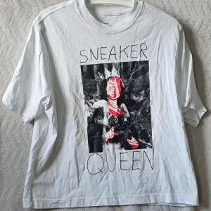 Vit Nike T-shirt Sneaker Queen - Vit T-shirt från Nike med trycket 'Sneaker Queen' och en cool grafisk bild med röda sneakers på framsidan. Modellen är loose fit och har korta ärmar. Perfekt för dig som älskar streetstyle och sneakers.
