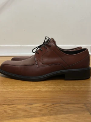 Bruna loafers från Ecco  - Säljer ett par stilrena bruna loafers i skinn med fyrkantig tå och svarta snören. Skorna har en låg sula i svart och klassisk design som passar till många olika outfits. Perfekta för dig som gillar en clean och tidlös look. Köpte de för min pappa men de passade inte