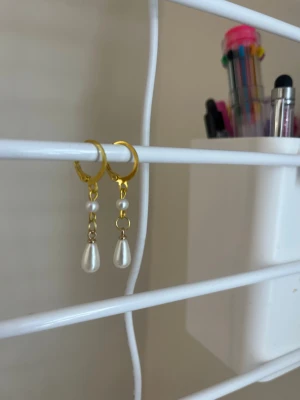 Guldiga örhängen med pärlor - Snygga örhängen med guldfärgade ringar och hängande vita pärlor. De har en droppformad pärla längst ner och en rund pärla ovanför, vilket ger ett elegant och trendigt uttryck. Perfekt för dig som gillar stilrena accessoarer med lite extra detalj. Handgjort 