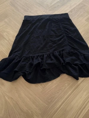 Svart kjol med volangkant - Svart kjol med asymmetrisk volangkant och lätt rynkade detaljer. Kjolen har en enkel design och är tillverkad i mjukt bomullstyg som känns bekvämt. Perfekt för dig som gillar en stilren look med lite extra edge.