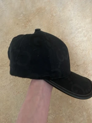 Svart keps från Gucci med GG-mönster - Snygg svart keps från Gucci med diskret GG-monogram över hela kepsen. Tillverkad i en mix av ull och kashmir för extra lyxig känsla. Klassisk böjd skärm och justerbar passform. Perfekt accessoar för dig som gillar exklusiv streetwear.
