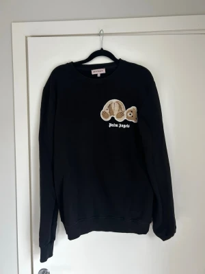 Svart sweatshirt från Palm Angels - Cool svart sweatshirt från Palm Angels med ikoniskt nallebjörnsmotiv på bröstet och vit logga under. Tröjan har rund halsringning och ribbade muddar vid ärmslut och nederkant. Perfekt för dig som gillar streetwear och vill sticka ut.