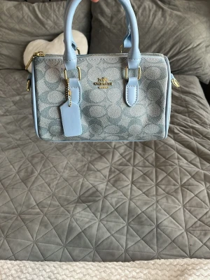 Ljusblå handväska från Coach - Säljer en snygg ljusblå handväska från Coach med klassiskt monogrammönster och guldfärgade detaljer. Väskan har både axelrem och handtag samt en tag med Coach-logga. Materialet är en mix av canvas och läder. Perfekt för dig som gillar stilrena accessoarer.