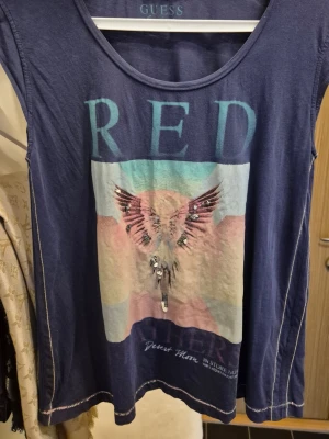 Blå topp från Guess med tryck - Cool blå topp från Guess med rund halsringning och tryck i pastellfärger på framsidan. Motivet har vingar och texten 'RED', dekorerad med glittriga detaljer. Perfekt för dig som gillar unika prints. Liten metallplatta med tryck Guess fastsatt i nedre kant.  Storlek XS i  VID modell