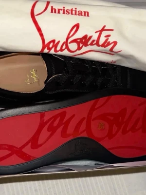 Christian Louboutin svarta sneakers - Säljer ett par svarta sneakers från Christian Louboutin med klassisk röd sula och guldfärgad logga på innersulan. Skorna har en låg profil, snörning och är tillverkade i ett lyxigt material som känns exklusivt och stilrent.