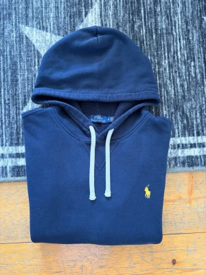 Mörkblå Polo Ralph Lauren Hoodie - Storlek M - Hej! 👋 Säljer en klassisk marinblå (Navy) Polo Ralph Lauren hoodie i storlek M. Den har den ikoniska pony-loggan broderad i gult på bröstet – en riktigt snygg kontrast! 🐎 Tröjan är i bra skick, välvårdad och knappt solblekt, vilket är ett stort plus för marinblått ✨ Den har kvar sin mjuka fleece-känsla på insidan och är 100% äkta med tvättlappar kvar ✅ Ingen QR-kod på denna modell. Ett stilrent premium-basplagg. Fraktar snabbt! 🚛📦💬💯 