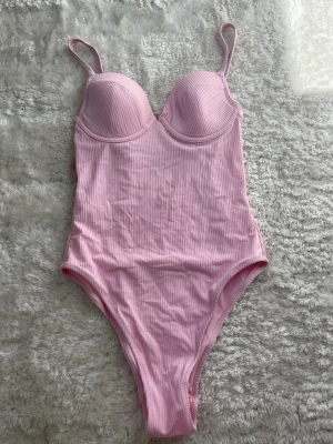 Rosa ribbad baddräkt  - Snygg ljusrosa baddräkt med tunna axelband och formade kupor. Materialet är ribbat och ger en cool struktur. Baddräkten har hög skärning vid benen och en feminin passform. Perfekt för strandhäng eller poolparty.